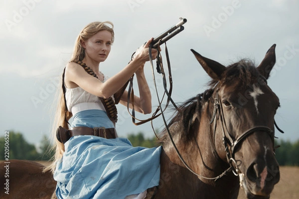 Fototapeta Portrait of a beautiful female cowgirl with shotgun from wild west riding a horse in the outback.