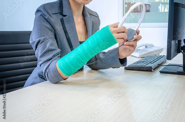 Fototapeta woman with broken hand and green cast  working on computer and holding headphone