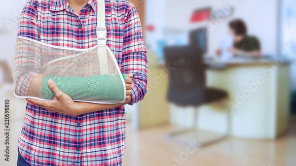 Fototapeta hand in a sling with broken arm in green cast isolated on blurred background working woman with office desk workplace in office