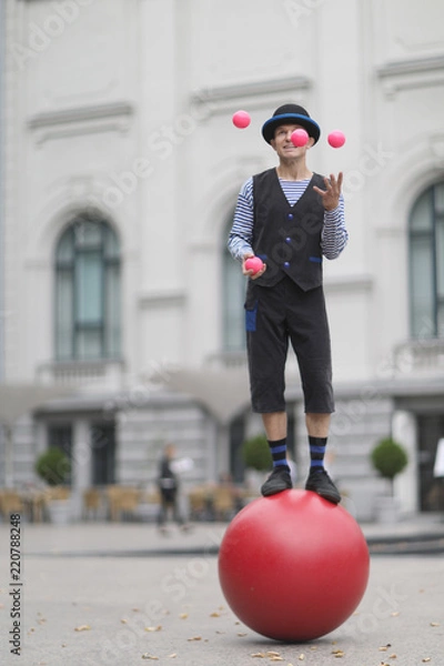 Fototapeta The clown juggles with pink balls, standing on a big red ball in the street of a European city