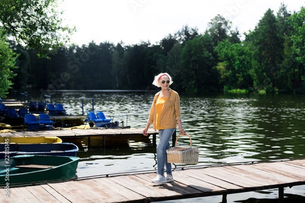 Fototapeta Woman in glasses. Elderly woman wearing glasses holding hamper for picnic in hand while crossing the bridge
