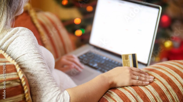 Fototapeta Closeup image of woman sitting next to Christmas tree and browsing online stores on laptop