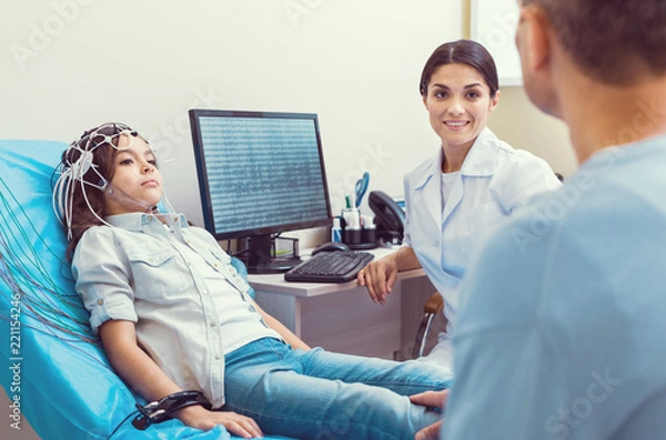 Obraz Stressed moment. Smiling female doctor smiling to a father while his poor little girl getting her brain analyzed by an electroencephalograph.