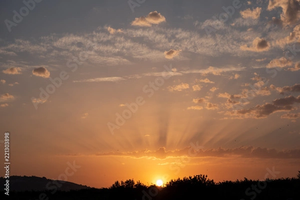 Fototapeta Sunset with sun almost hidden and shadows on the clouds and blue sky