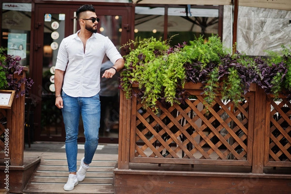 Fototapeta Stylish tall arabian man model in white shirt, jeans and sunglasses posed at street of city. Beard attractive arab guy.
