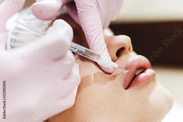 Obraz The process of lips enhancement. Cosmetician makes an injection of hyaluronic acid in a beautiful lips. The young girl with a beautiful face in the special hat and hands of the doctor in the pink glov