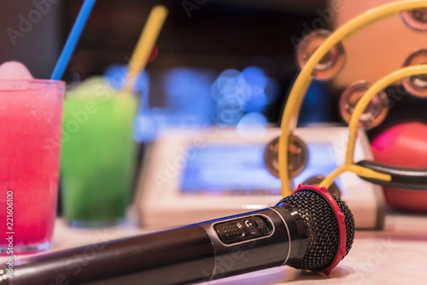 Fototapeta Czarny mikrofon w klubie karaoke, z pilotem, napoje gazowane melon i truskawka, żółty tamburyn i ekran do śpiewania muzyki na scenie.