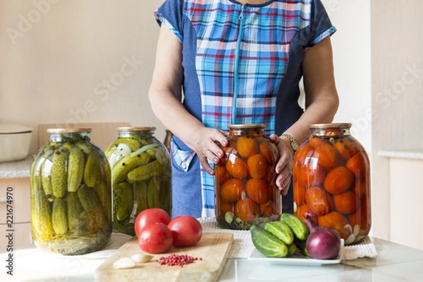 Fototapeta woman in the kitchen next to the table. on the table are banks with pickled, salted tomatoes and cucumbers, on the table fresh tomatoes, cucumbers, onions, garlic, vegetables