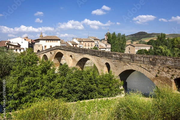 Fototapeta Spain, Puente La Reina, Gares: Panoramic view of famous romanesque bridge over river Arga with skyline of Spanish small town literally named 'Bridge of the Queen', green riverside and blue sky.