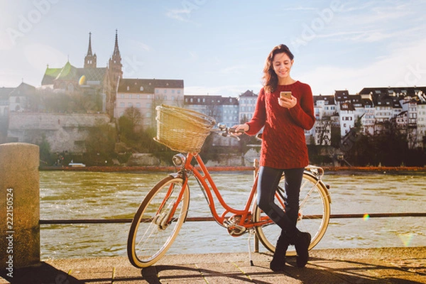 Fototapeta theme tourism on bicycle and modern technology. Beautiful young caucasian woman stands near red retro bicycle on riverside river Rhine Basel Swiss winter warm sunny weather uses the phone in hand.