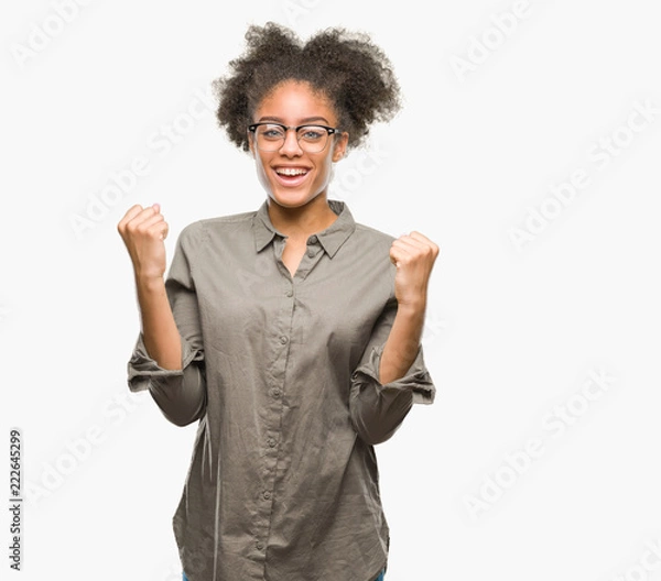 Fototapeta Young afro american woman wearing glasses over isolated background celebrating surprised and amazed for success with arms raised and open eyes. Winner concept.