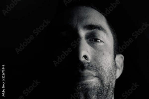 Obraz Dramatic portrait of a young man half expressionless face in deep shadow looking at camera isolated on black background