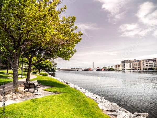 Obraz Beautiful landscape scenery shot along the Fox River just after sunrise in Green Bay Wiscosin with riverwalk trail, bench, trees, grass and rocky shoreline framing the shot and blue cloud filled sky.