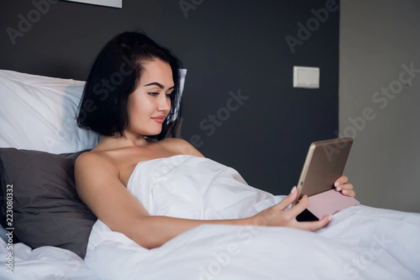 Fototapeta Portrait of happy young Caucasian woman sitting in bed, using digital tablet and smiling