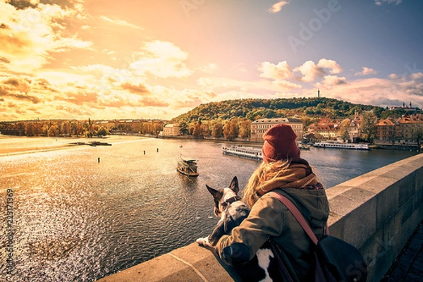 Obraz Young women tourist with a puppy dog and a backpack looking at the tourist boat and swans sailing on Vltava river from the Charles Bridge (Karluv Most) in Prague, Czech Republic