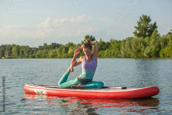 Fototapeta Young woman doing yoga on sup board with paddle. Meditative pose, side view - concept of harmony with the nature