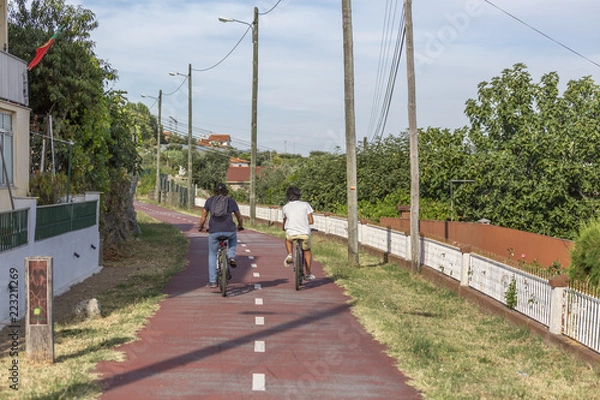 Fototapeta Two boys cycling on pedestrian cycle path, with street lamps, blue sky as background and vegetation, in Viseu, Portugal