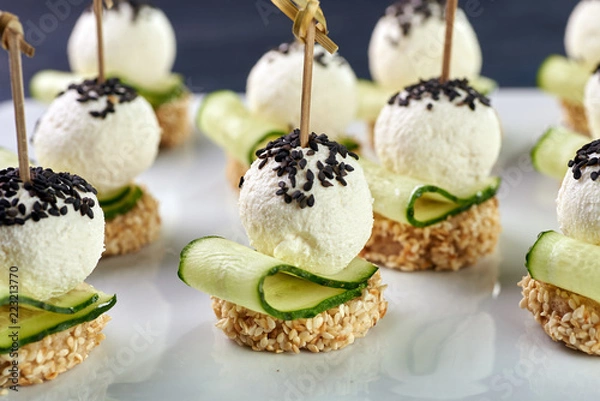 Fototapeta An unusual beautiful appetizing appetizer for a festive table. Mini canapes from a piece of baguette with sesame, thin slice of cucumber and a ball of cheese. Exotic black sesame for decoration.