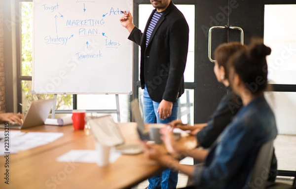 Fototapeta Photo of Business partners brainstoming and discussing documents about merketing plan at meeting. Selective focus at wording "Marketing Plan" on whiteboard.