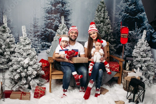Fototapeta Studio portrait of cheerful caucasian family in Santa hats with kids sitting on their knees holding Christmas presents with snowed fir trees around them. Looking at camera and smiling happily.