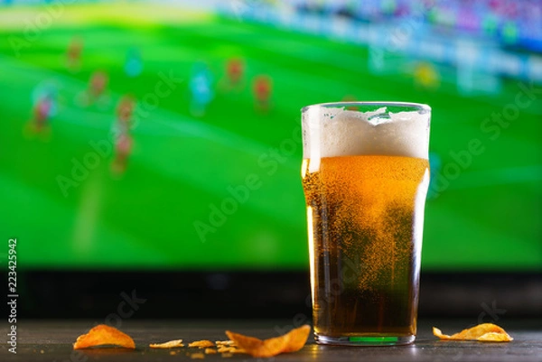 Fototapeta A glass of beer on a dark wooden table with crushed potato chips. Football on a background, high resolution