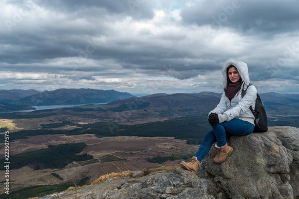 Obraz Woman with fluffy white jacket and hiking shoes posing on a mountain with mountains and lakes in the backgroud, on a cloudy day at Nevis Range, Scottish Highlands