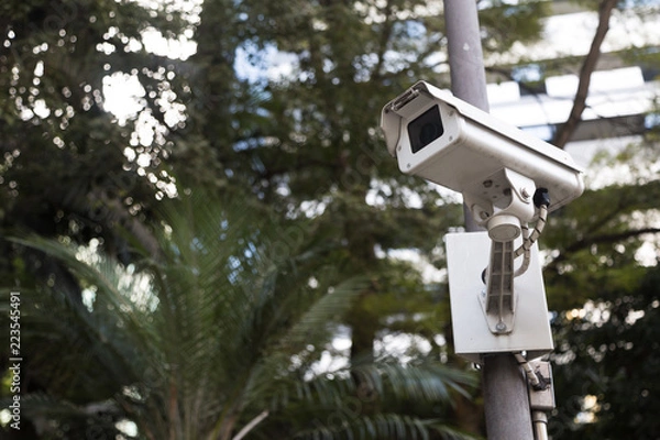 Fototapeta Bellow view of a security camera(cctv) in a public place. With a metal camera, green trees and buildings in the background we compose this photo. Secure, systens, technology and monitoring as concept
