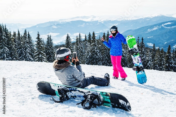 Fototapeta two women in ski suits on top of the mountain do selfie. Girls with snowboards sit on the snow and are photographed