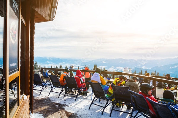 Fototapeta skiers sit on chairs near a restaurant on top of a mountain. People in ski suits rest and drink