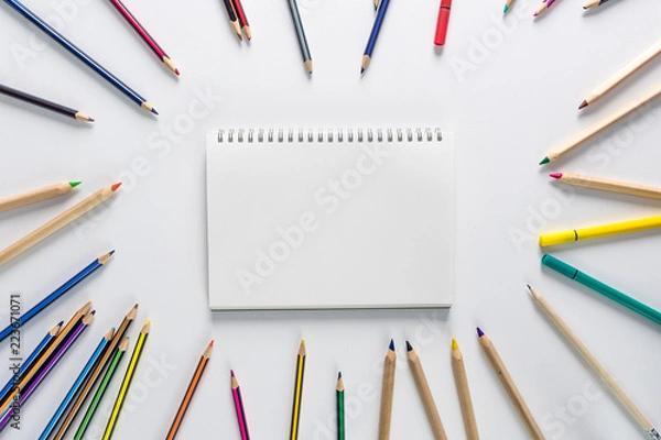 Obraz Top view over a school supplies as coloured pencils placed in a circle on a white background with blank work book in the middle of it. Back to school concept. Office supplies flat lay