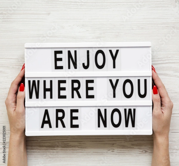 Fototapeta Female hands hold modern board with 'Enjoy where you are now' word over white wooden background, top view. Business concept. From above, flat-lay, overhead.