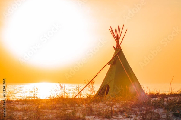 Fototapeta Wigwam on a sandy seashore. American native indian building outdoors in summer. Teepee on a sundown, in warm sunlight against the sea