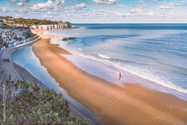Fototapeta Low tide at Stone Bay, Broadstairs, Kent as summer turns to autumn, a lone surfer walks on the beach and a family on the promenade along side the beach huts and white cliffs.