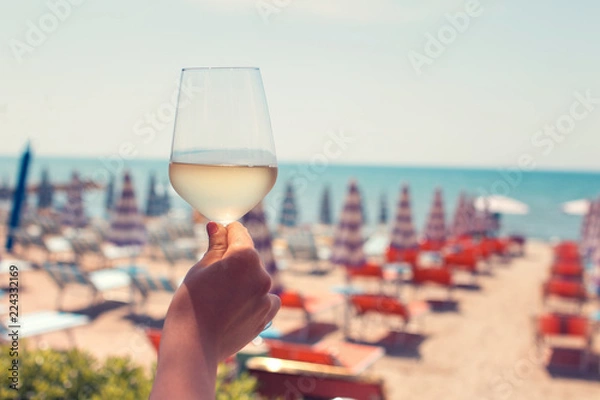 Fototapeta Female hand with a glass of white wine on the background of a beautiful beach. Travel vacations concept. Toned