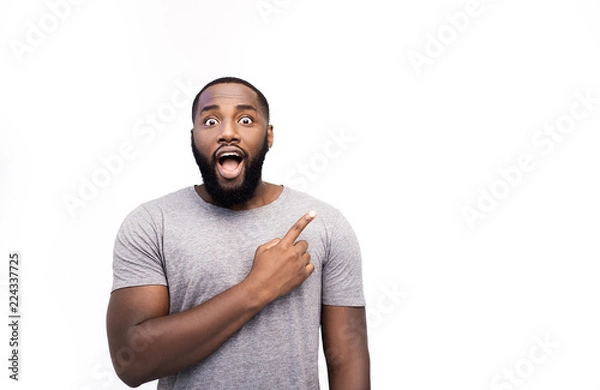 Fototapeta African American man grins at camera, indicates copy space for tex or logo, advertises something. Surprised man points with fore fiinger, shocked expression, isolated over white background. Look here!