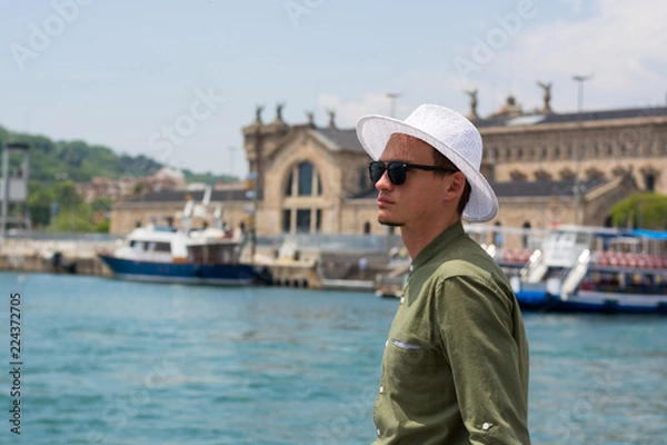 Fototapeta A young guy in a hat, sunglasses stands in the port of Barcelona's at the port customs background, Barcelona, Spain