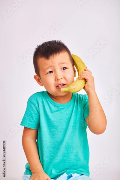 Fototapeta A child boy in a turquoise shirt, holds a banana in place of a phone while talking about a conversation - fruits and a healthy diet