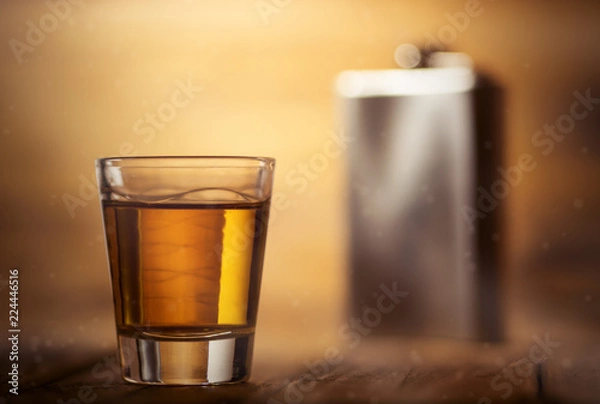 Fototapeta Whiskey shot glass on top of a wooden table background and a metal hip flask in the background