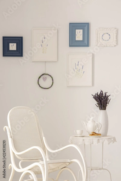 Fototapeta Flowers on table next to armchair in white living room interior with posters on the wall. Real photo