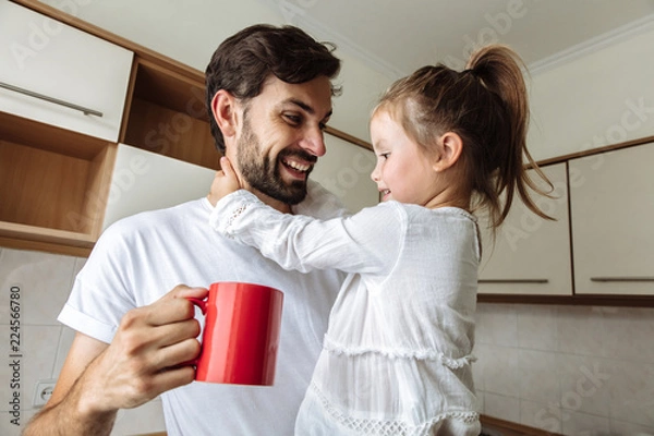 Obraz Parenthood. Family. Home. Dad is holding a cup and his little daughter in his arms. Both are talking and smiling. In the kitchen at home