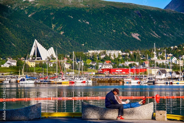 Fototapeta A couple sits on a bench in front of the calm, picturesque harbor at Tromso in far northern Norway in summer, with colorful boats and the Arctic Cathedral in the background