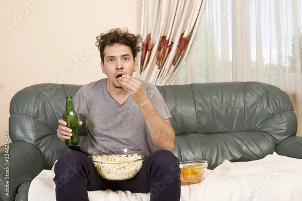 Fototapeta Podekscytowany młody człowiek z pilotem i popcornem na kanapie przed telewizorem