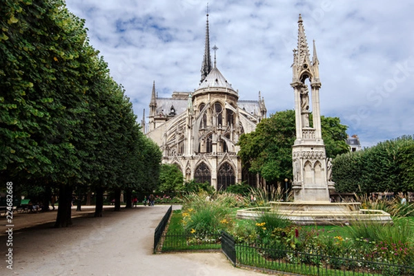 Fototapeta Paris, France - August 13, 2017. Medieval Notre Dame de Paris, most visited french monument of Gothic architecture. View from Square Jean XXIII - park with Virgin Fountain and flowers near Cathedral.