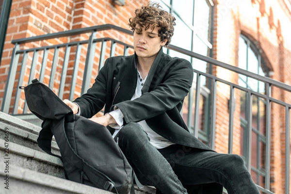 Obraz Young attractive pensive man sitting on steps and rummaging in his backpack.