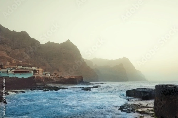 Obraz typical evening island scene with hazy dusty cliffs and the crystal clear blue sea with the buildings of a small village