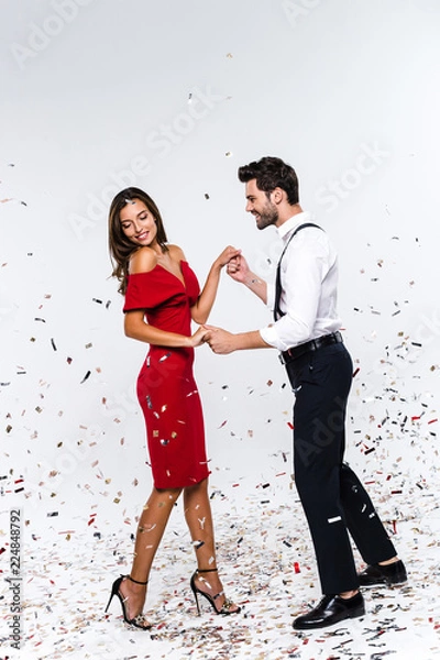Fototapeta Perfect celebration for two. Full length of young beautiful couple dancing while standing against white background with confetti 