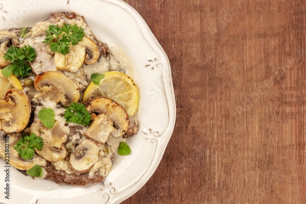 Fototapeta Overhead closeup photo of scallopine di vitello, veal scallopini, with a mushroom sauce, lemons, and parsley, on a dark rustic texture with copy space