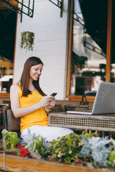 Obraz Happy smiling girl in outdoors street coffee shop cafe sitting at table with laptop pc computer, texting message on mobile phone friend, in restaurant during free time. Mobile office freelance concept
