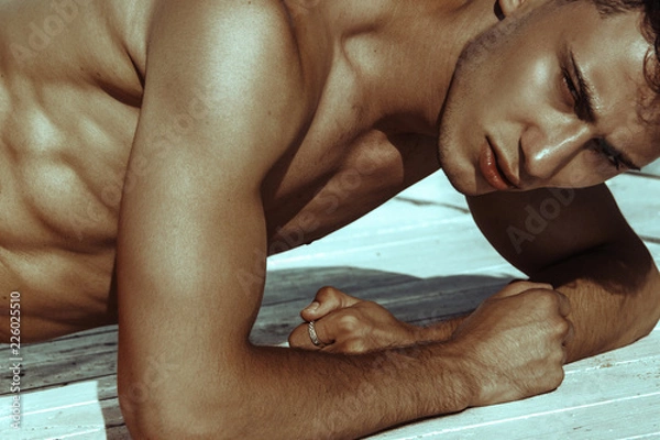 Obraz Strong face muscle male model in the open  summer shower on the vacation pool background with blue water on the nature beach on the sunset