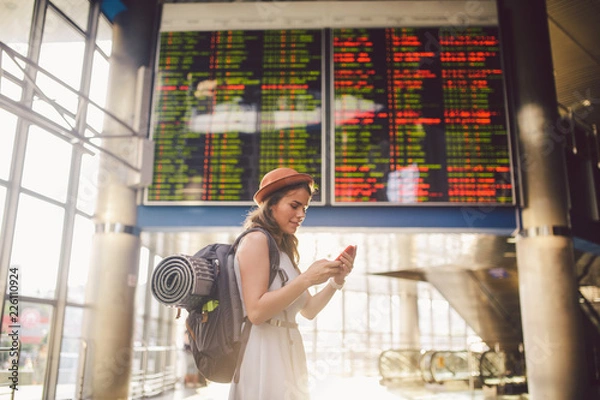 Fototapeta Theme travel and tranosport. Beautiful young caucasian woman in dress and backpack standing inside train station or terminal looking at a schedule holding a red phone, uses communication technology